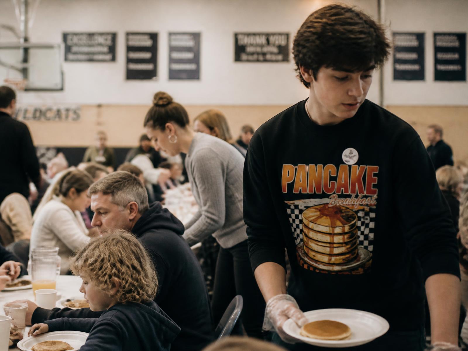 Teen volunteer in a printed community sweatshirt carrying plates at a school pancake breakfast